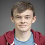 James Devine's headshot. A middle aged white man with short brown hair smiling at the camera. He is wearing a blue shirt and a red hoodie.